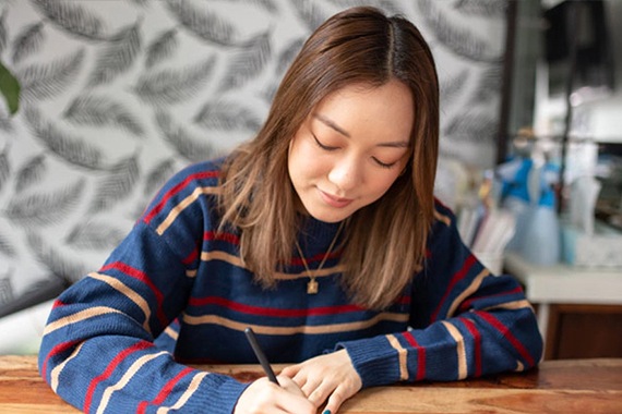 Amanda Rejč Li kreira bulet planer na svom Galaxy Tab S6 Lite pomoću tamnosive S Pen olovke dok sedi za drvenim stolom. Nosi mornarsko plavi džemper sa crvenim i roze prugama.