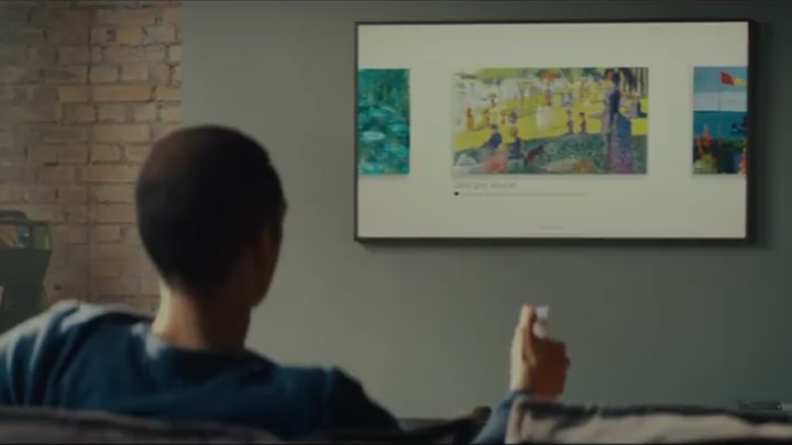 A man is using a remote control to browse some paintings that are shown on The Frame TV that is mounted on his living room wall.