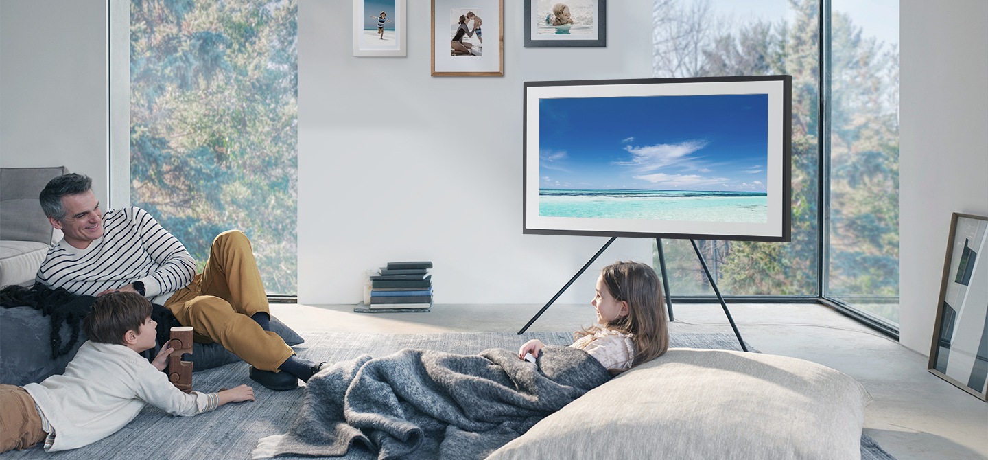 رجل وطفلاه يلعبون لعبة بجوار The Frame. يوجد تلفزيون The Frame على حامل أرضي ويعرض صورة زرقاء لامعة لمياه الشاطئ تحت سماء زرقاء صافية.