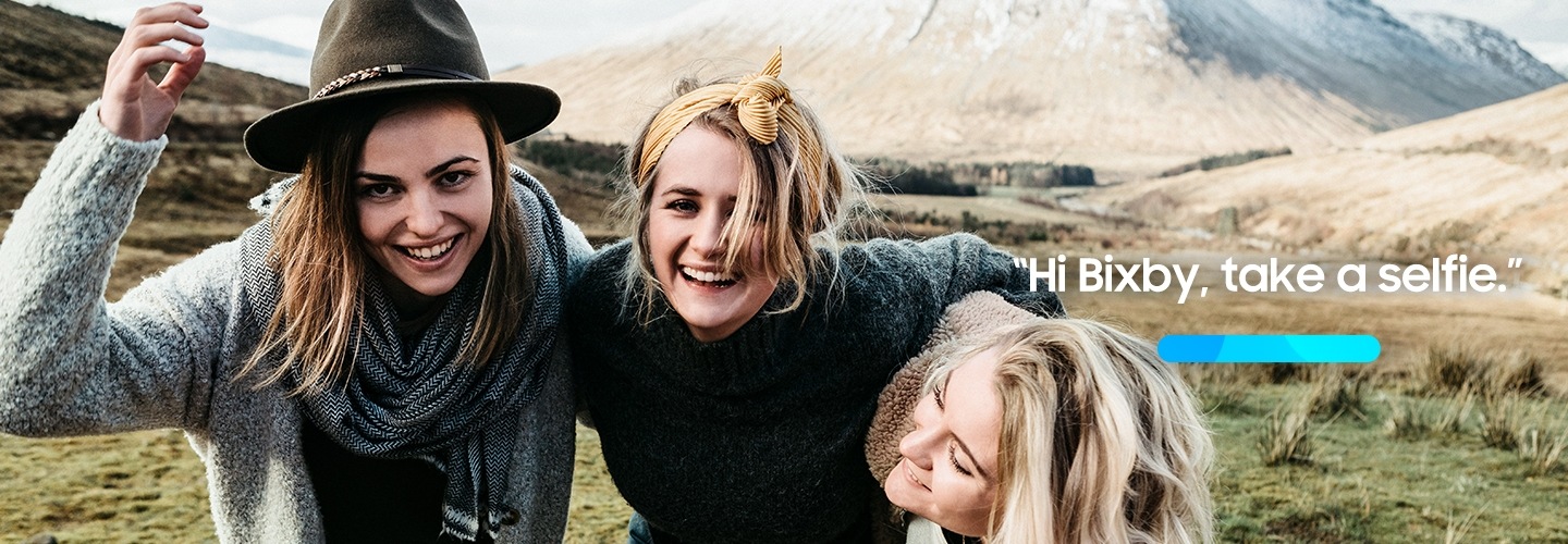 Three women standing on the mountain, putting arms around eath other's shoulders and laughing together. A progress bar icon and text "Hi Bixby, take a selfie" are shown.