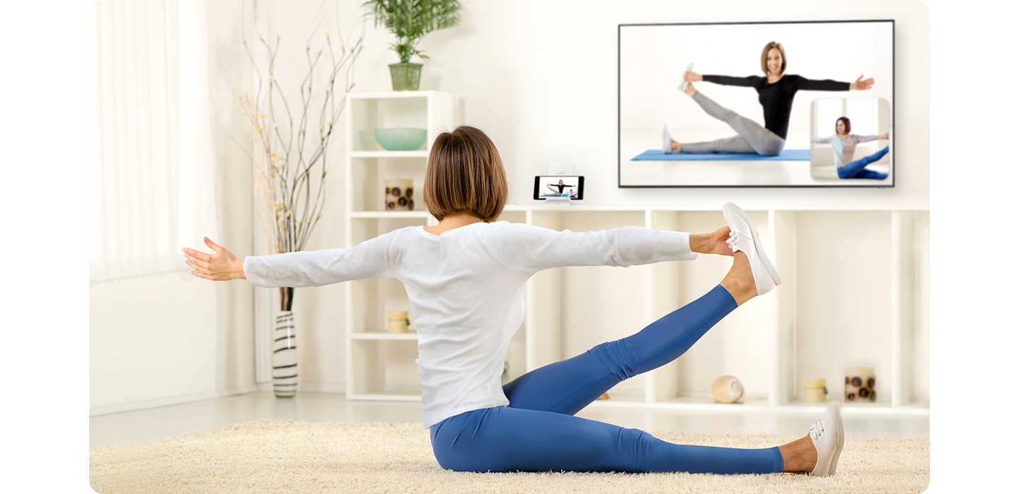 A woman, sitting down in her living room, is following a yoga pose showing on the TV in front of her. Next to the TV, her smartphone is recording her pose, which is being mirrored onto a small window on the lower righthand corner of her TV so that she can compare her pose to the instructional video playing.
