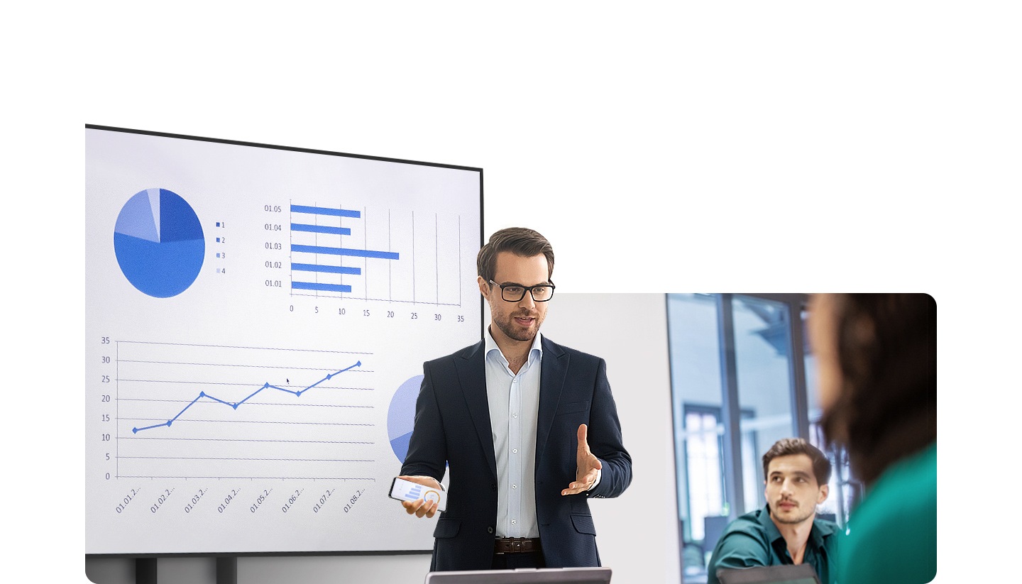A man is giving a presentation in front of people in an office. Behind him, a large display is showing various graphs and he is holding his Galaxy smartphone in his hand.