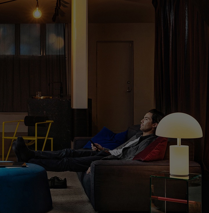 A man in a dimly-lit living room sitting back on a sofa with his feet up on an ottoman. He watches a tv show with remote control in hand.