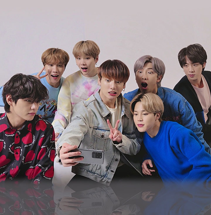 BTS taking Group Selfie. All members are sitting on the ground and taking a BTS selfie on a Galaxy S20+ held by Jung Kook.