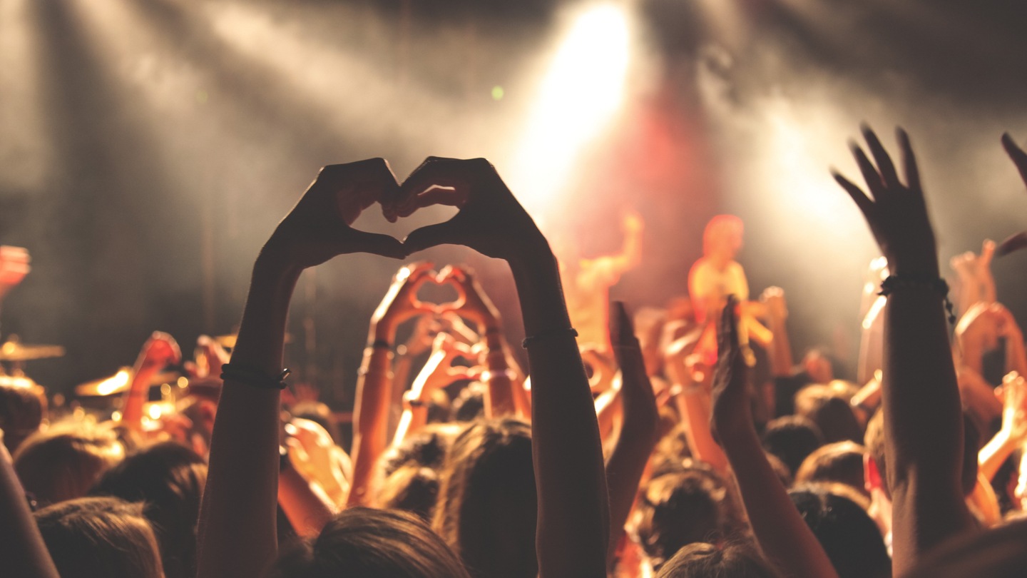 Concert scene at a dark venue with flashing spotlights. There is a large audience in front of a stage where a band member is playing the guitar. There are people in the audience raising their arms and forming a heart shape with their hands.