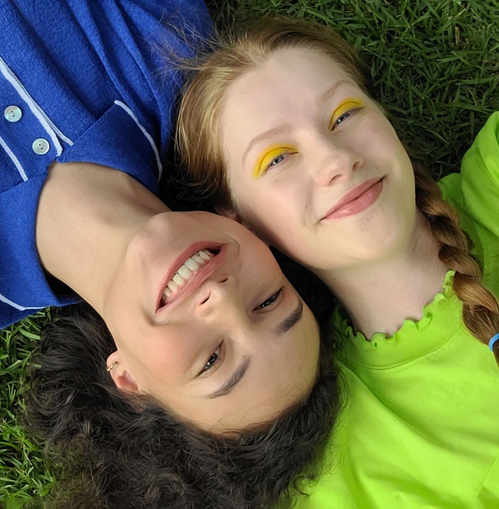 An aerial view of two women laying side-by-side on the grass. The woman on the left is wearing a blue collared shirt while the woman on the right is wearing a lime green top. Their heads are pressed together from opposite ends. They're both smiling at the camera.