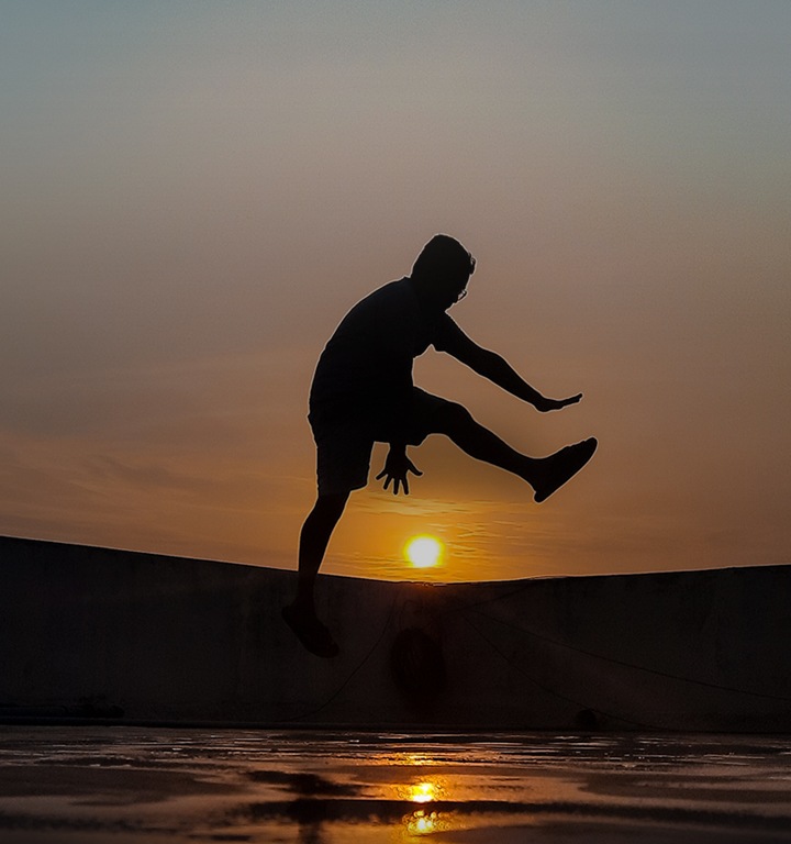 kv A young male at the beach pretends to jump over the setting sun in the background.