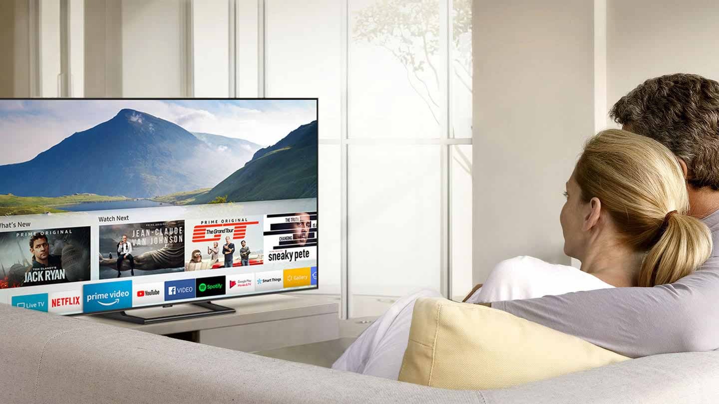 "2 people are watching Smart TV and Smart Hub UI is displayed over a nature background image on TV screen; The UI contains various content, such as, 'What's New', 'Watch Next', and several applications like Netflix, Amazon Prime Video, YouTube, Facebook Video, Google Play Movies & TV, SmartThings, Gallery, Internet.