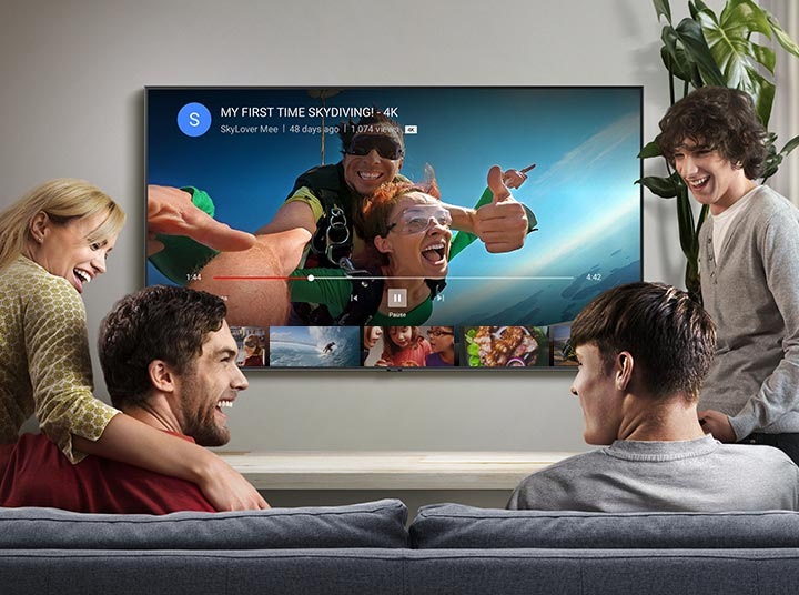 Fire personer sitter i stuen og ser kvalitetsvideoer fra YouTube på Samsung Smart TV. Video av fallskjermhopping med 4K-logoen blir strømmet gjennom YouTube-appen på Smart TV.