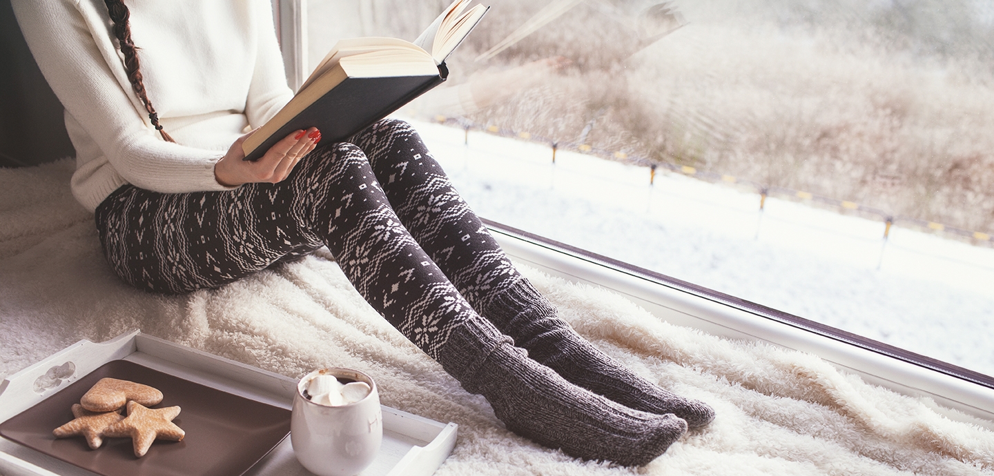 니트와 겨울바지, 두꺼운 양말을 신은 여자가 바닥에 앉아 책을 보고 있습니다.