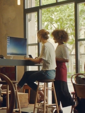 한 여자가 컴퓨터 앞에 앉아 있고, 한 여자는 옆에 서 있습니다.