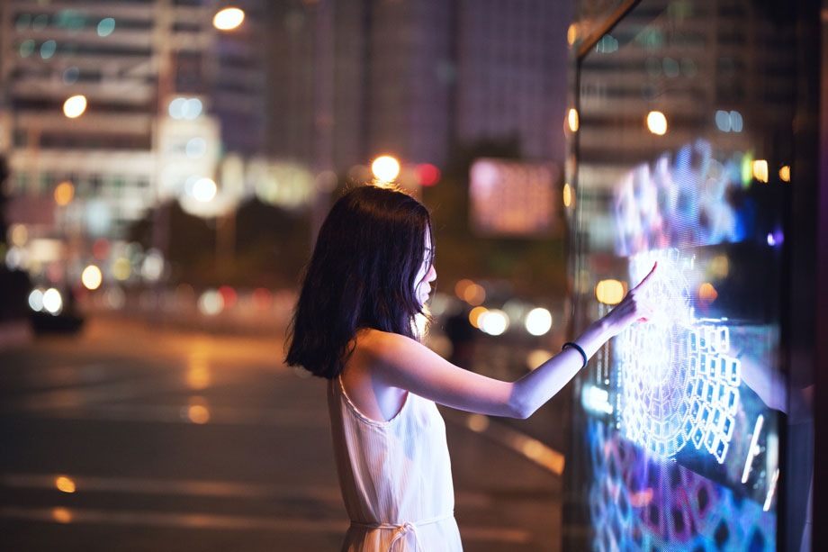 조명이 빛나는 거리 위에 서 있는 소녀가 현란한 색감의 터치 스크린 디스플레이를 터치하고 있습니다.