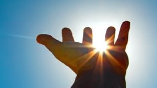 Hand reaching up toward the sky, with the sun seen between its fingers.