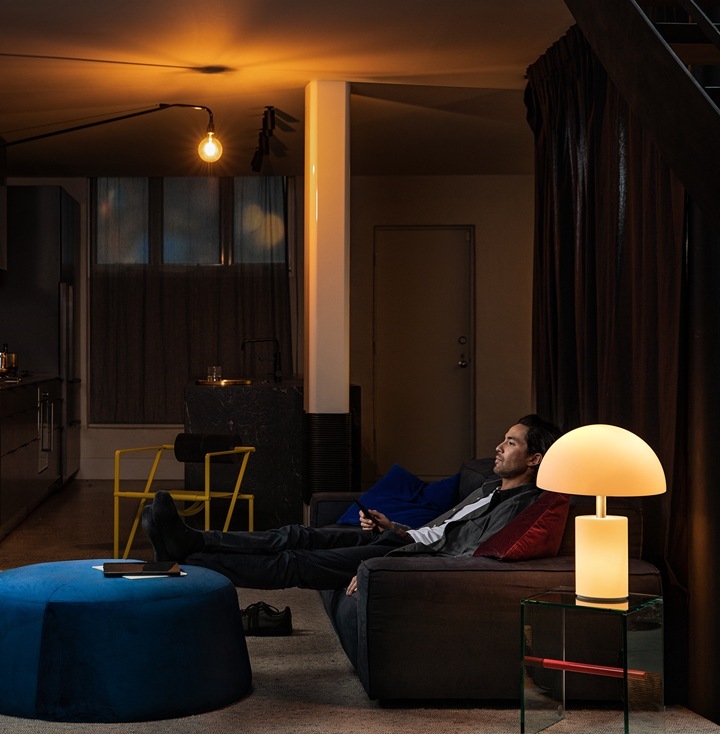 A man in a dimly-lit living room sitting back on a sofa with his feet up on an ottoman. He watches a tv show with remote control in hand.
