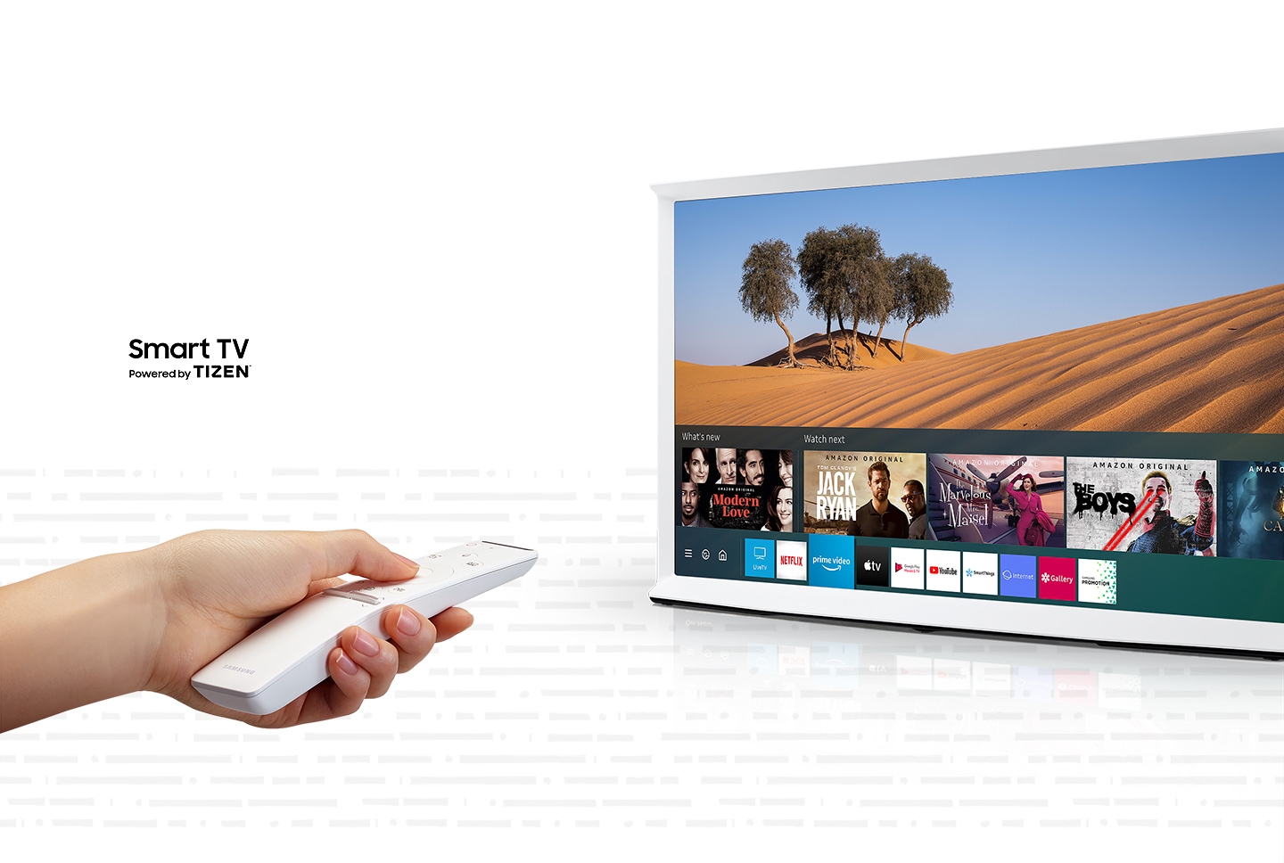 The screen of a Cloud White model of The Serif shows a desert scene with the SmartHub open at the bottom. To the left, a hand is holding a white Samsung One Remote, pointed at the TV. The Serif TV is Smart TV powered by Tizen.