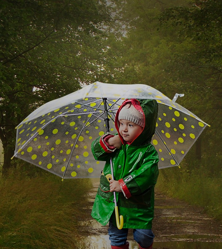 A boy in a green raincoat is holding yellow polka dot umbrella and walking along a stand of trees.