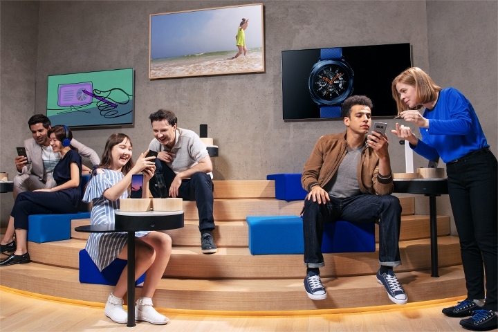Five customers seated on steps inside a store, in two groups of two along with a single man receiving smartphone guidance from a Galaxy Consultant
