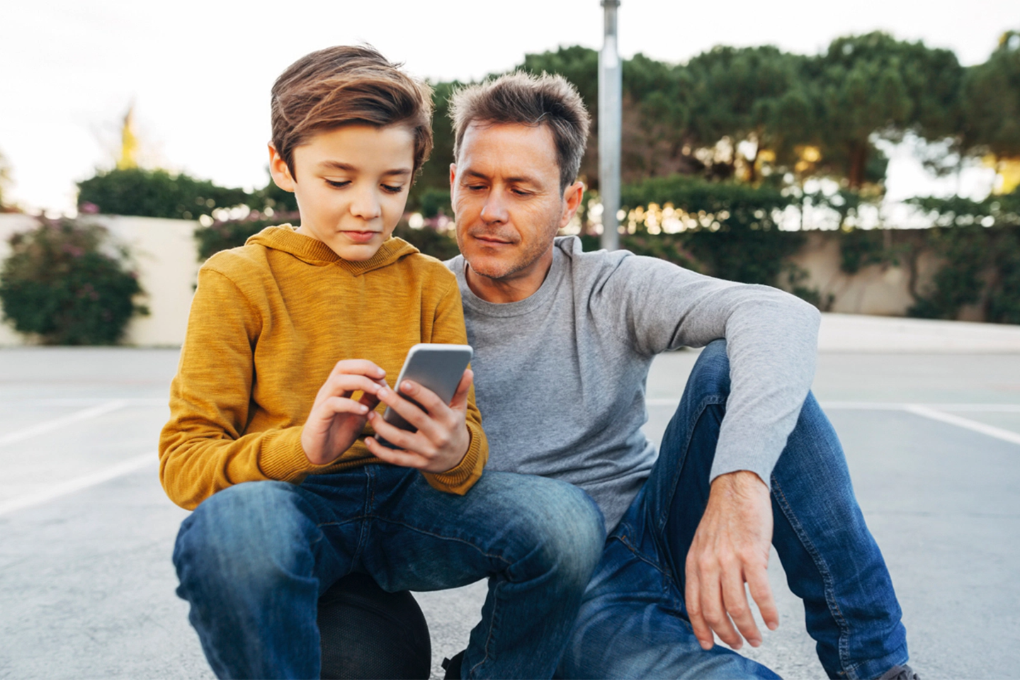 Çocuğunuza telefon alırken dikkat etmeniz gereken 5 önemli nokta Bir basketbol topunun üzerinde oturan ve yanında oturan babasıyla birlikte telefonuna bakan küçük bir çocuğu gösteren bir açık hava sahnesi.