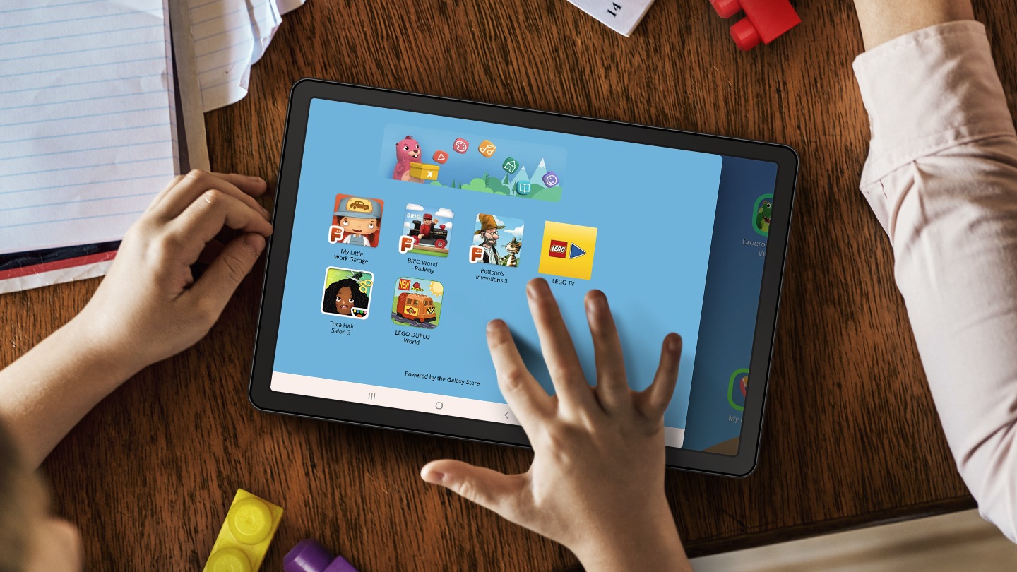 Bir masanın üzerinde çevresinde kağıtlar ve lego parçaları olan bir tablet. Çocuğun elleri yakında ve sağ eli tabletin ekranına dokunuyor. Ekran üzerindeki LEGO Uygulaması kullanıcı arayüzü, çocuğunuzun tecrübe edebileceği farklı eğlence türlerini gösteriyor.