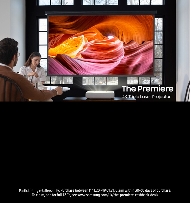 A man & woman sit in a modern living room with a Samsung The Premier 4K laser projector projecting photos from a wooden table A man & woman sit in a modern living room with a Samsung The Premier 4K laser projector projecting photos from a wooden table