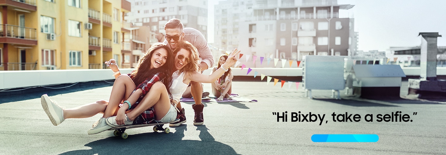 2 women sit on a skateboard and a man crouches behind them. A progress bar icon and text "Hi Bixby, take a selfie" are shown.