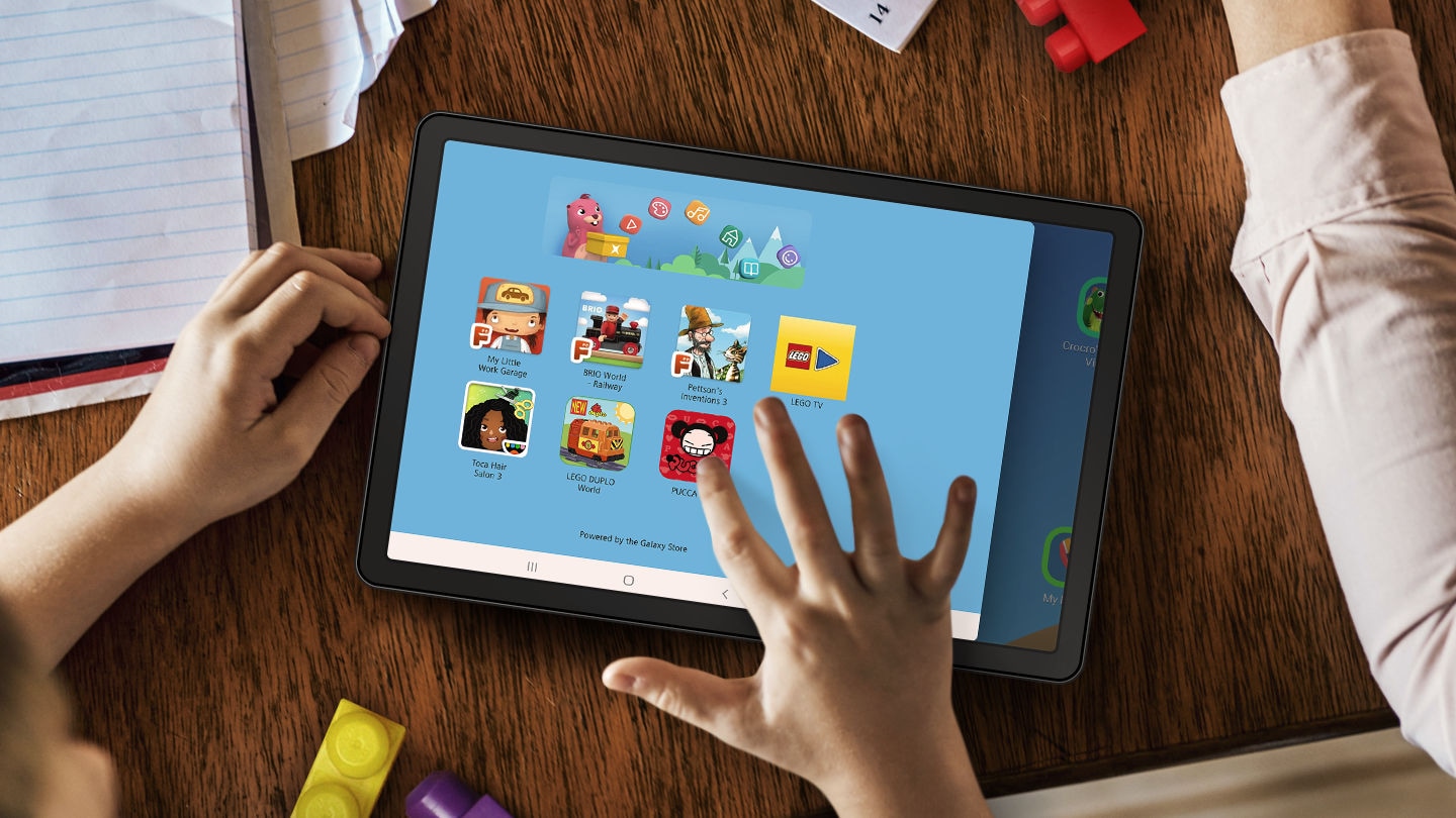 A tablet on a table surrounded by papers and LEGO blocks. A child’s hands are nearby and their right hand is touching the tablet screen. Onscreen is the LEGO App UI, showing different kinds of entertainment your child can experience.