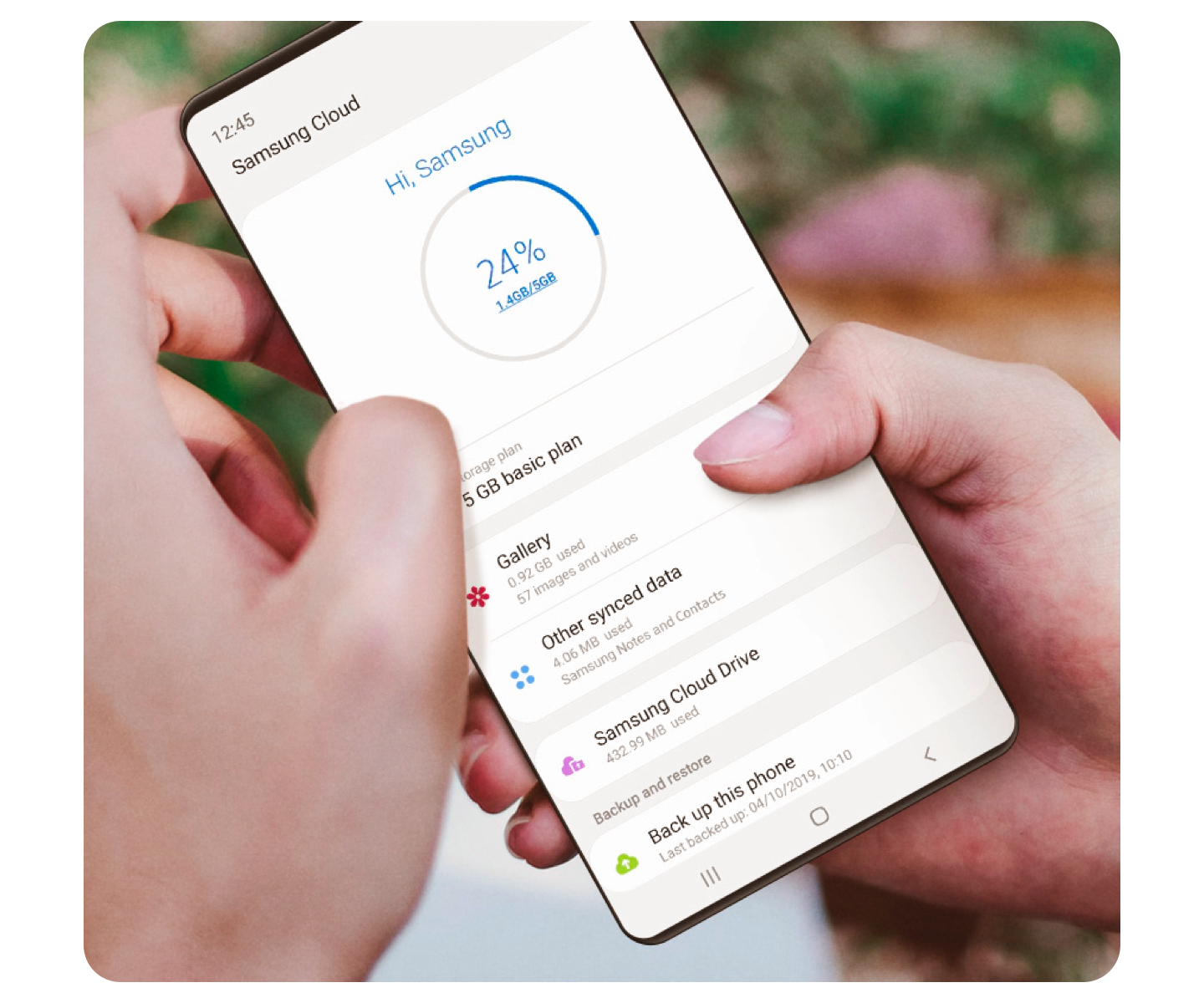 Two hands hold a Samsung smartphone with Samsung Cloud application open on the screen with a green blurry background behind