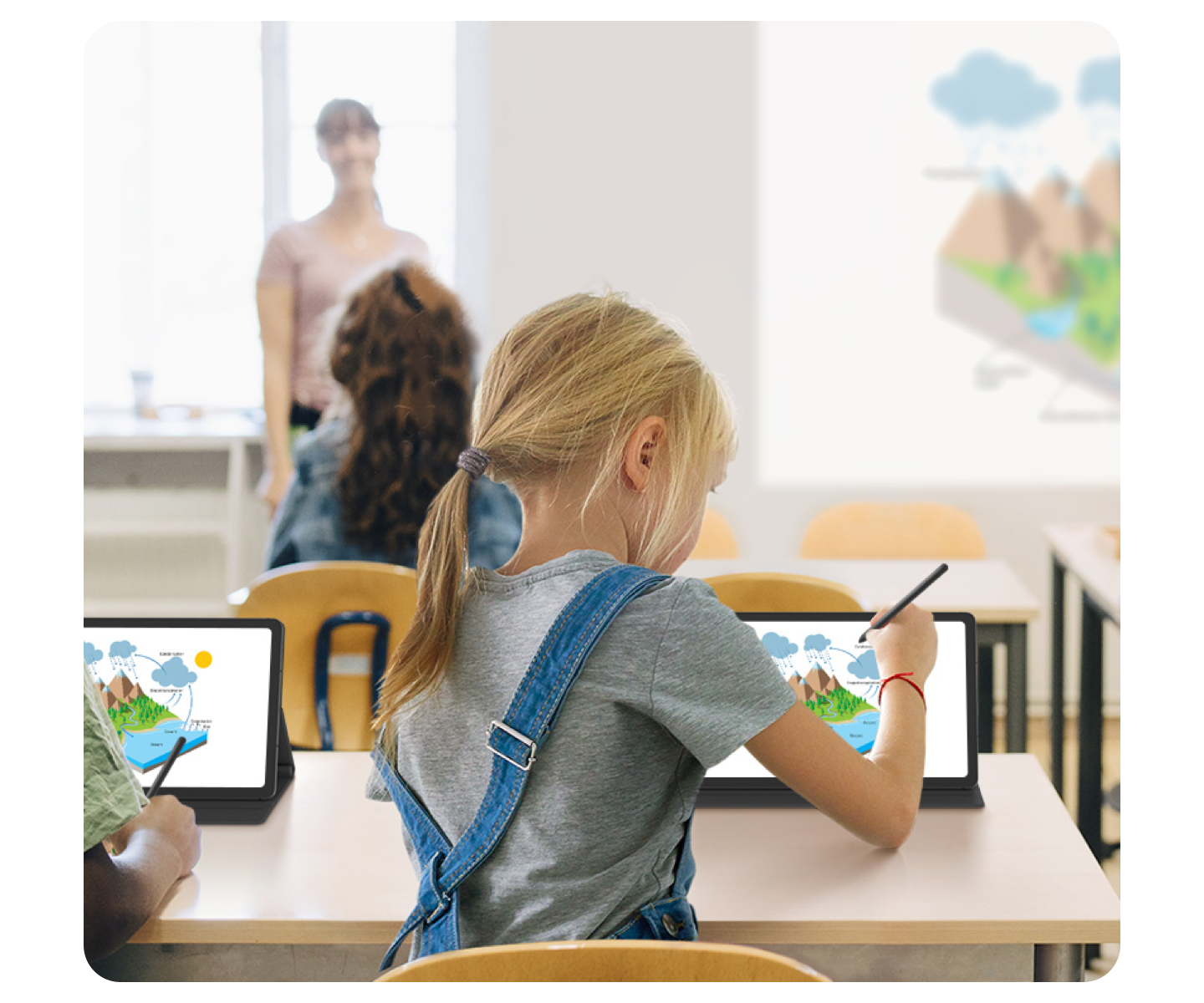 A young girl sits at a desk in a bright classroom, drawing on a black Samsung tablet using an S Pen. A blurred out teacher stands at the front.