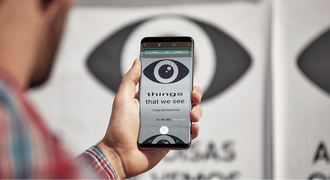 Fotografia unei persoane care ține în mână un telefon Galaxy S9+ cu un semn de traducere pe ecran și fotografia netradusă în fundal.