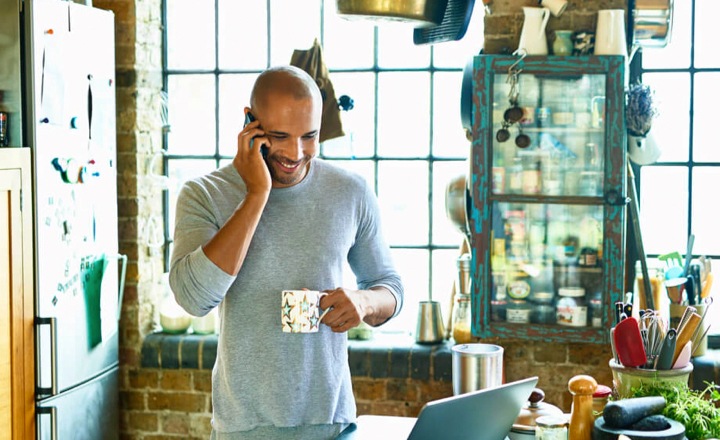Front view of a man standing in a kitchen with coffee mug in his hand. He is talking on a phone representing Samsung smartphones as gift for Father's day