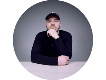Headshot of Lewis Hilsenteger against a gray background. He's wearing a baseball cap and long-sleeve shirt, sitting at a table with his hand on his chin