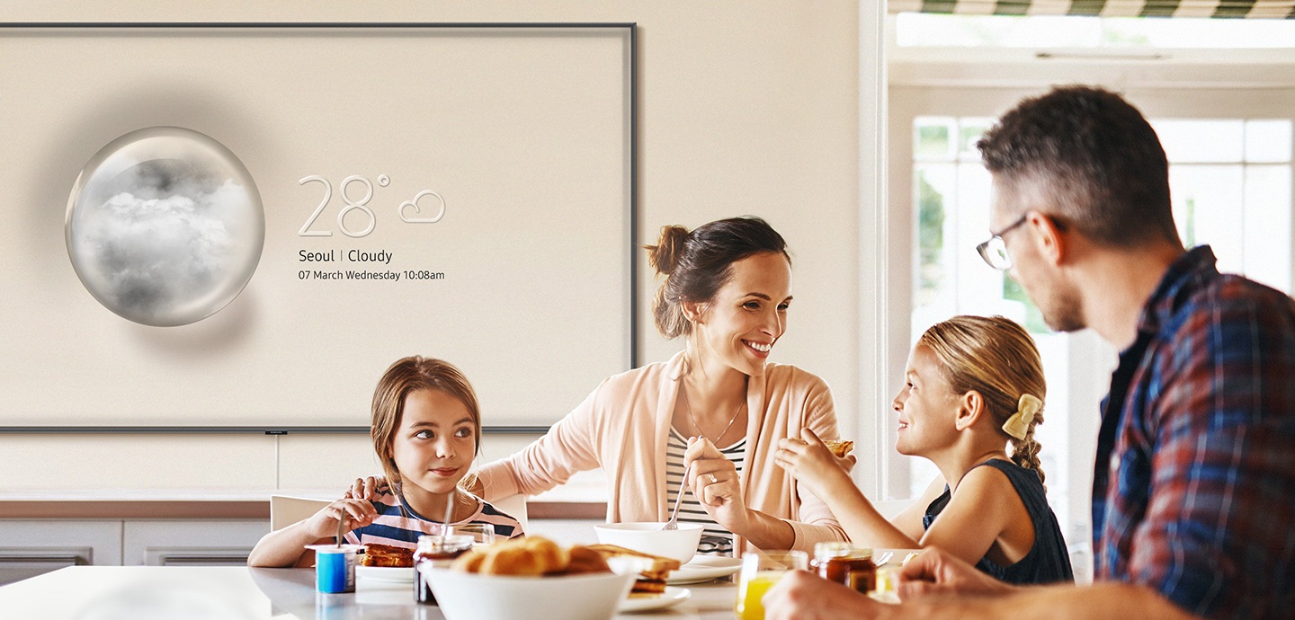 Una familia está comiendo sentada alrededor de una mesa. Detrás de ellos, el televisor QLED se encuentra colgado en la pared con el modo Información activado. Se muestra el modo Información y el televisor QLED con el modo Ambiente cambia al panel de información del clima en la cocina.