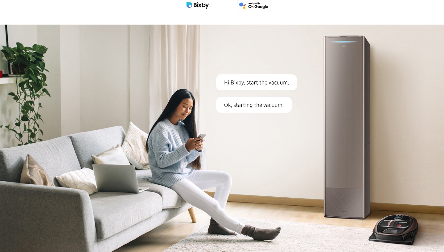 A woman sits on her sofa with her smartphone. There is a smart air conditioner and a smart robot vacuum in the room. 2 conversation bubbles appear near the woman with the text "Hi Bixby, start the vacuum" and "Ok, starting the vacuum". 3 AI assistant logos are displayed above: Bixby, Hey Google, and Amazon Alexa.