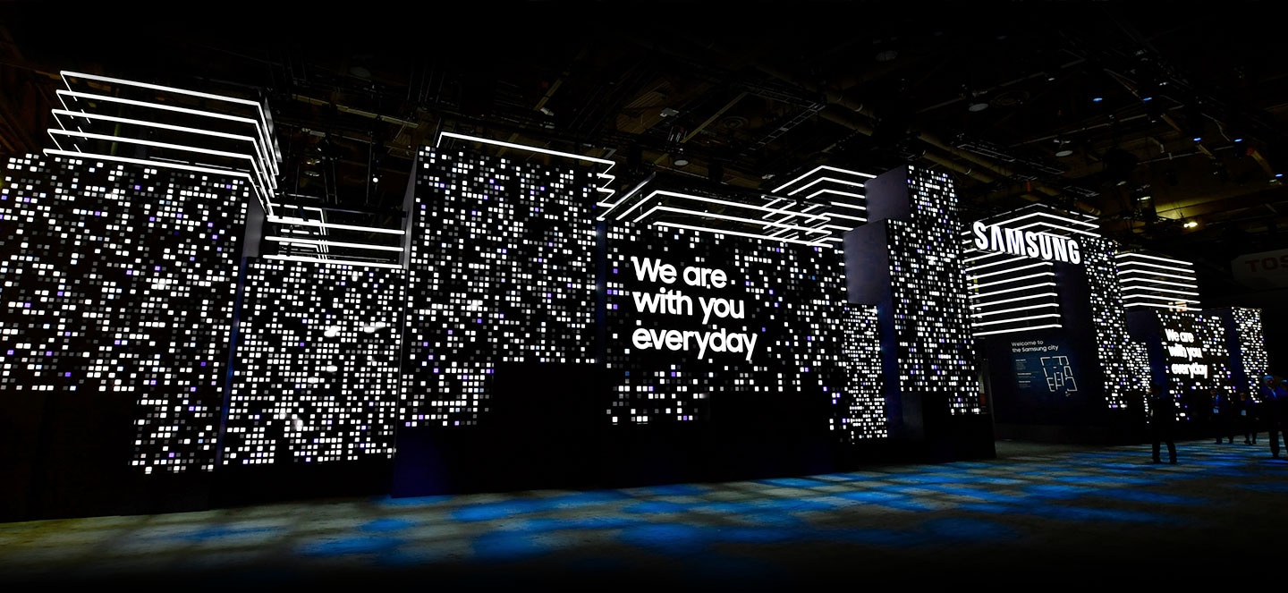 A large LED display is lit up to mimic an urban backdrop in line with the theme of Samsung City with the words "We are with you everyday" featured prominently in the center. The floor below the display is lit up with lights in shades of blue and yellow.