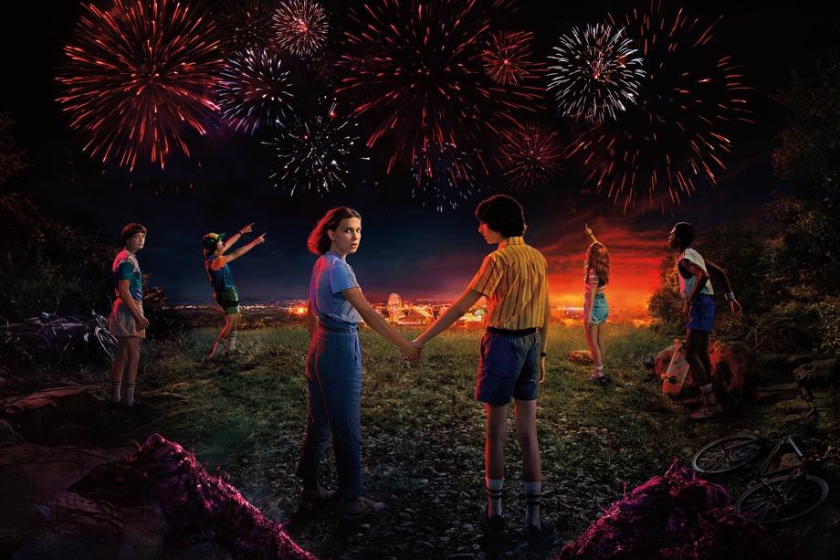 Netflix 모바일 앱의 인기 시리즈인 <기묘한 이야기>의 사진입니다. 들판 위에 서 있는 일레븐과 마이크가 손을 잡고 있고 다른 아이들은 불꽃놀이를 바라보고 있습니다.
