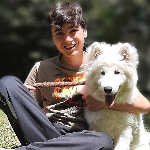 A boy with one arm around a white dog, hugging, while they are sitting on grass