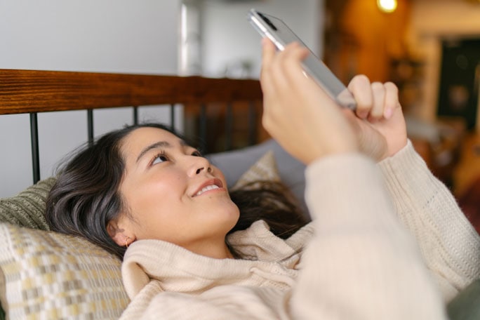 한 여성이 침대에 누워 스마트폰을 들고 화면을 보며 웃고 있습니다. 
