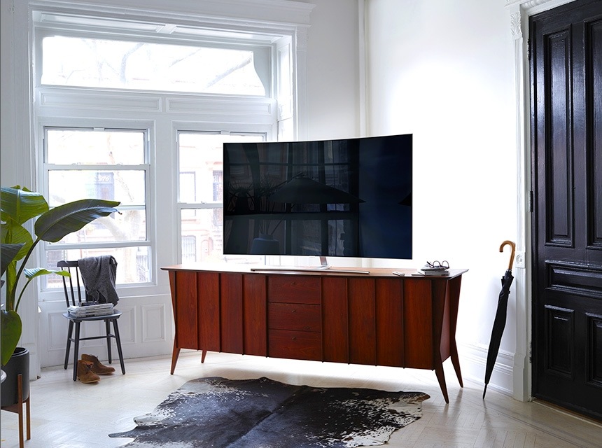 Bàn gỗ cứng được đặt ở góc trong phòng khách tông màu trắng với TV QLED. Tấm thảm được đặt trước bàn với ghế và bình hoa bên trái và dù bên phải.