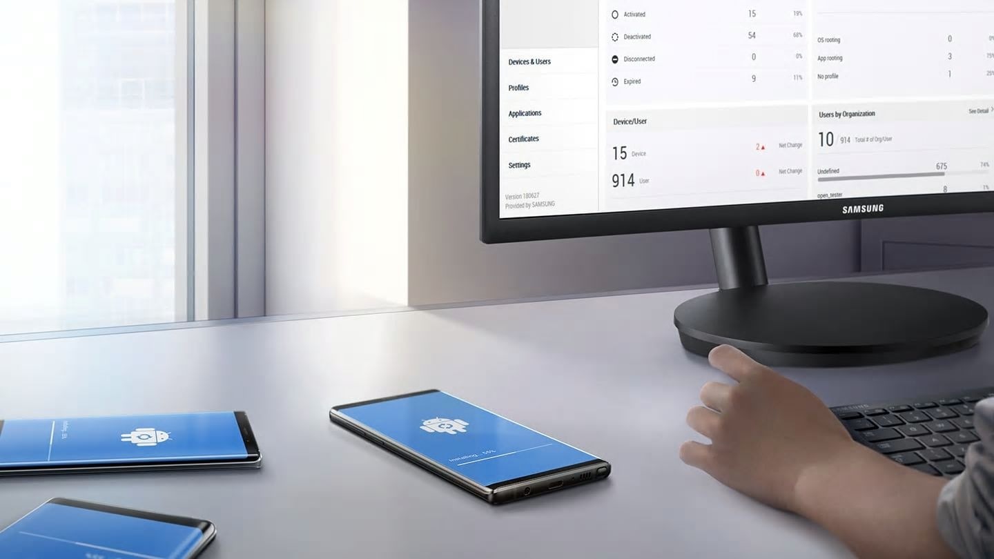 Image of a person sitting at a desk with multiple Galaxy Note9s displaying the Android update GUI and the Knox Manage GUI displayed on the monitor