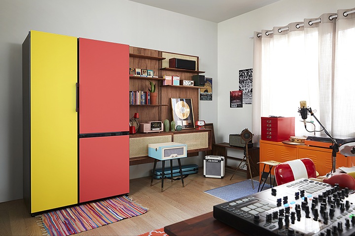 A BESPOKE refrigerator with bright yellow and red doors stands in a colorfully decorated living room filled with retro-style furniture