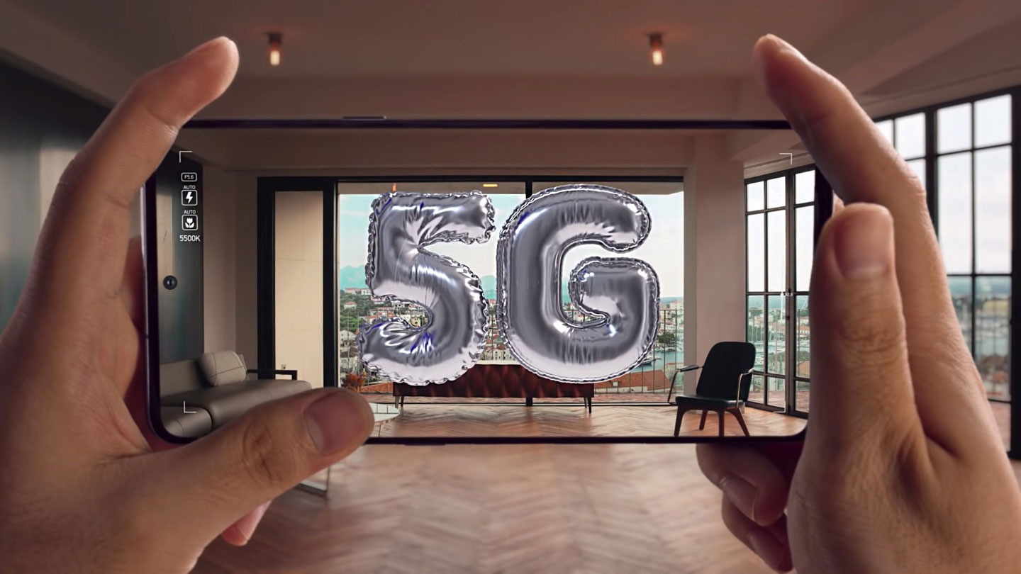 Two hands hold up a Galaxy device taking a picture of 5G balloons in a stylish, high rise apartment.