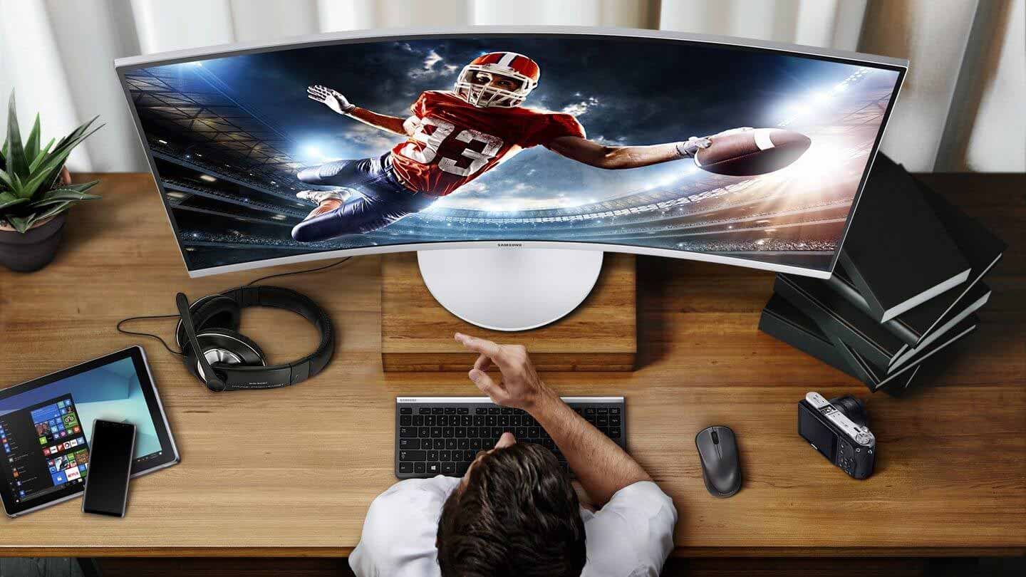 There is a monitor on the room table, and rugby sport is shown on the monitor screen. The man is staring at the monitor, and on the table are tablets, cell phones, headphones, mice, keyboards, cameras, potted plants, and books.