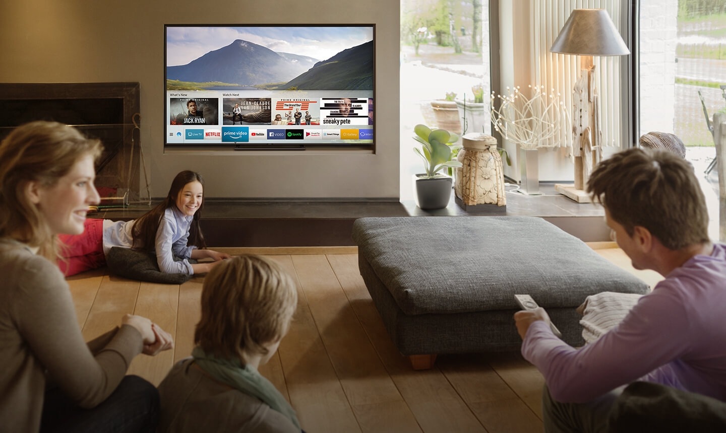 People are watching Smart TV in the living room and Smart Hub UI is displayed over a nature background image on TV screen; The UI contains various content, such as, 'What's New', 'Watch Next', and several applications like Netflix, Amazon Prime Video, YouTube, Facebook Video, Google Play Movies & TV, SmartThings, Gallery, Internet.