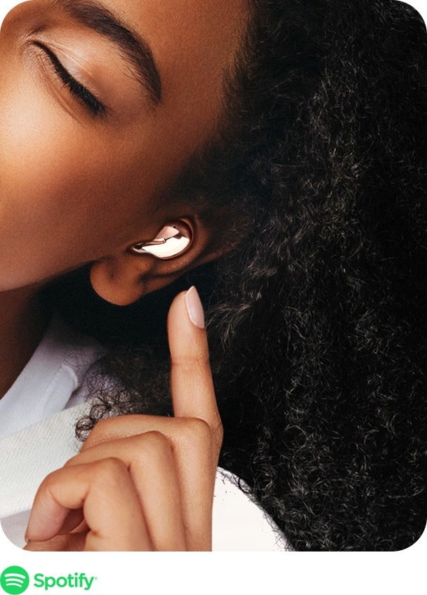 Kobieta ze słuchawkami bezprzewodowymi Galaxy Buds Live w kolorze Mystic Bronze korzysta ze Spotify, dotykając słuchawki Galaxy Buds Live. Poniżej widać Galaxy Note20 z graficznym interfejsem użytkownika Spotify.