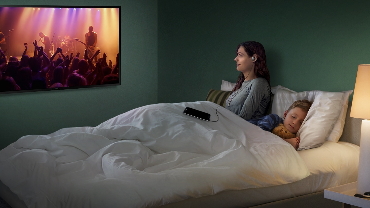 침실의 모습이 보입니다. 침대 위에는 곰인형을 안고 자고 있는 남자 아이의 모습과 벽면에 기대어 이어폰을 들으며 TV를 시청하고 있는 여자의 모습이 보입니다. TV 화면에는 공연장의 모습이 보입니다. 