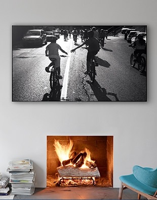 거실에 작은 의자가 있고 의자 앞에는 벽난로가 보여집니다. 벽난로 위에는 제품이 걸려있으며 화면 안에는 아이들이 자전거 타는 모습이 보여지고 있습니다.