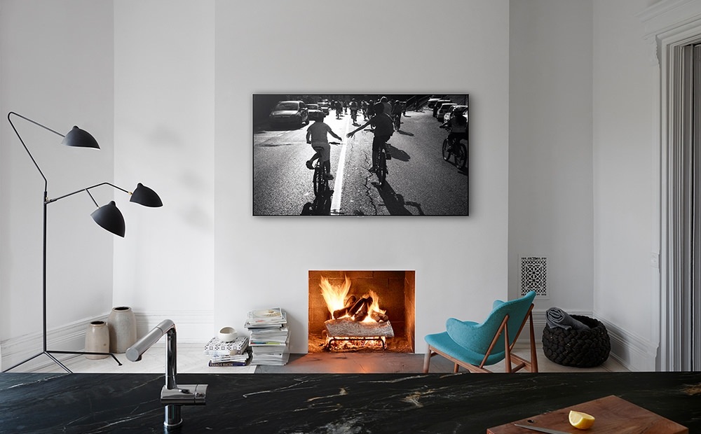 거실에 작은 의자가 있고 의자 앞에는 벽난로가 보여집니다. 벽난로 위에는 제품이 걸려있으며 화면 안에는 아이들이 자전거 타는 모습이 보여지고 있습니다.