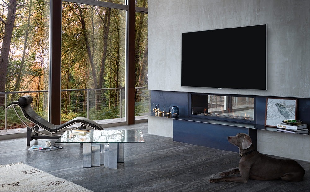 창밖이 훤희 보이는 거실에 쇼파와 검정색 강아지가 있습니다. 쇼파 앞에는 벽에 제품이 걸려있으며 제품하단에는 작은 선반이 있습니다.