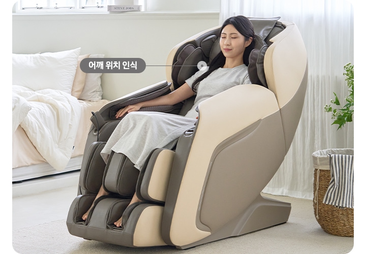 화이트톤 침실이 보이며 왼쪽에는 침대가 보입니다. 침대 옆에는 코지마 안마의자를 사용하고 있는 여성이 보입니다. 어깨 부분에 어깨 위치 인식이라는 텍스트가 보이며 화살표로 이어져 있습니다.