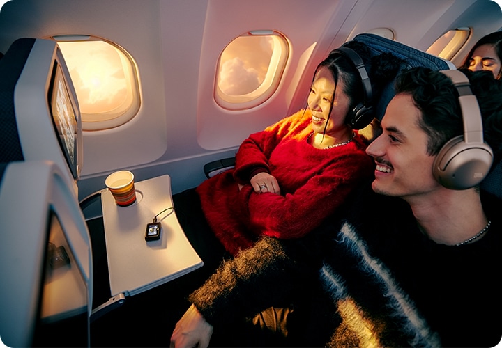 헤드폰을 착용한 남녀가 비행기 기내 엔터테인먼트 시스템에 트랜스미터를 연결하여 영상을 보고 있습니다.